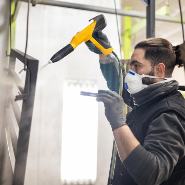 Un gros plan d'un ouvrier en tenue de protection et gants utilisant un pistolet pulvérisateur pour appliquer un revêtement noir sur une pièce métallique détaillée dans un atelier de fabrication.